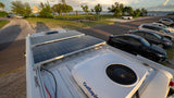 Top-down view of an RV rental roof with two solar panels and an OutEquip AC unit, parked by a waterfront scenic park.