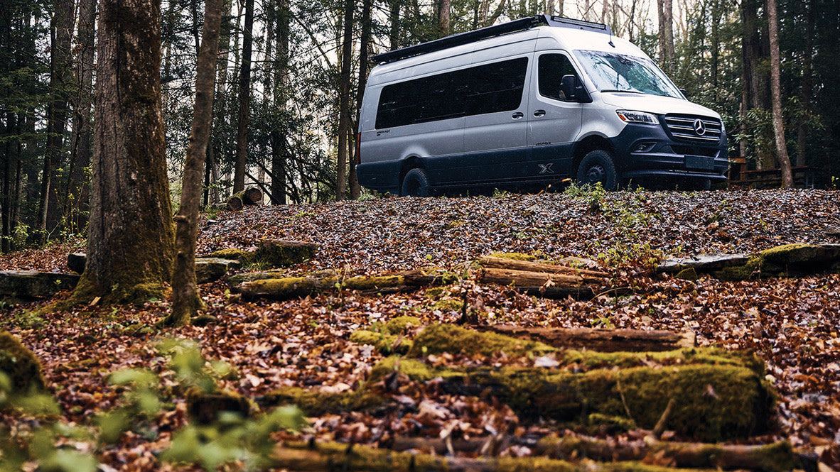 Silver Mercedes Sprinter camper van on a leaf-covered forest hill, ideal for an RV rental outdoor adventure.