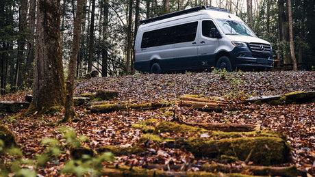 Silver Mercedes Sprinter camper van on a leaf-covered forest hill, ideal for an RV rental outdoor adventure.