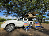 White truck with a rooftop tent & camp setup under a large tree. A perfect outdoor RV rental for shaded adventure.