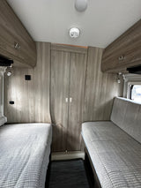 RV rental interior: light wood flooring and cabinetry, plush gray seating, a clear aisle, and safety devices. A modern camper living space.