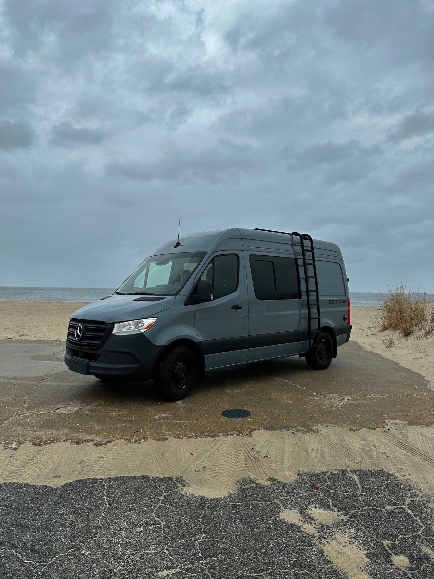 Grey Mercedes Sprinter van, ideal for RV rental, parked on a wet beach path under a cloudy sky.