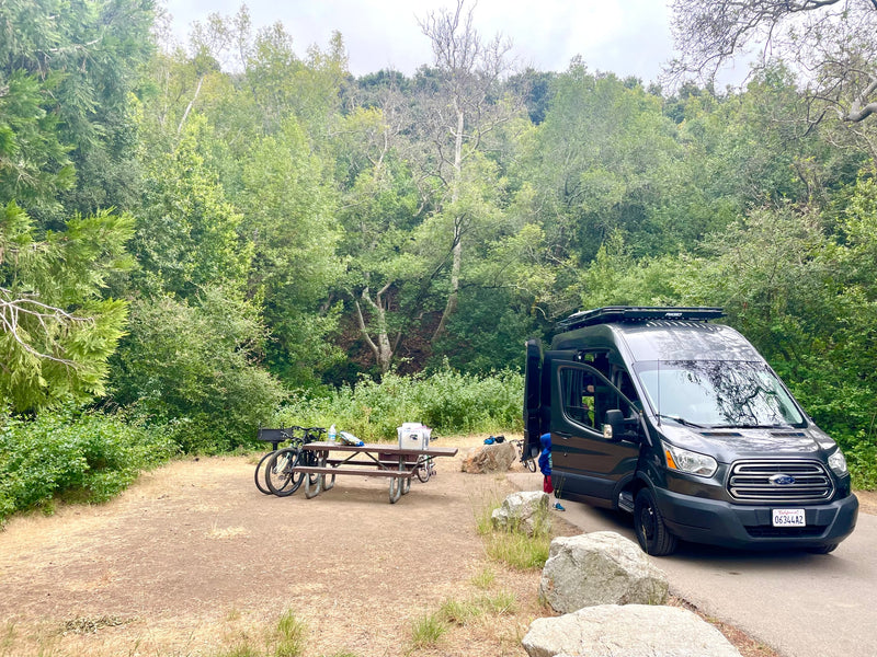 RV rental camper van parked at a green campsite with a picnic table, grill, and bicycles, surrounded by lush trees. — photo 39