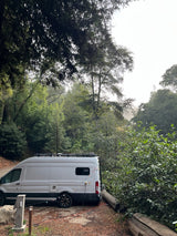 This rv rental camper van is set up for a cozy stay in a lush, green forest campsite, complete with string lights.