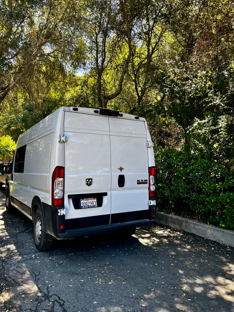 White Ram ProMaster camper van, great for rv rental, parked on asphalt with sunlit trees in the background.