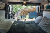 Cozy RV rental interior: A comfortable bed with pillows and shelving, offering a serene view out the back of the camper van.
