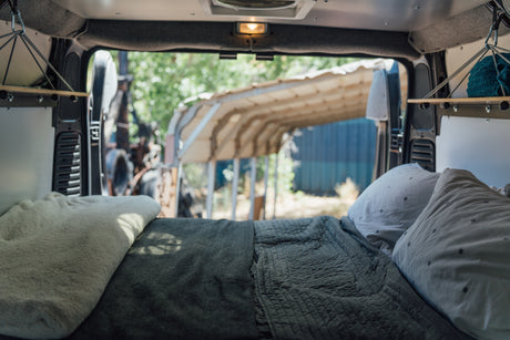 Cozy RV rental interior: A comfortable bed with pillows and shelving, offering a serene view out the back of the camper van.