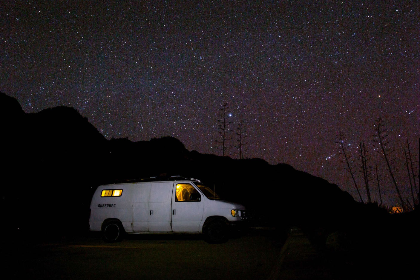 A white RV rental van, lit from within, parked under a brilliant star-filled night sky and dark mountains.