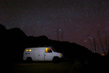 A white RV rental van, lit from within, parked under a brilliant star-filled night sky and dark mountains.