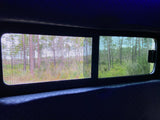 Inside an RV rental, two windows frame a pine forest view; one window has a screen, the other offers a clear glimpse.
