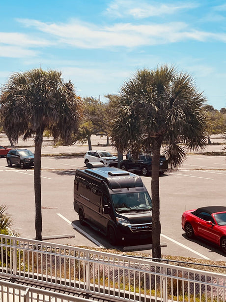 Black RV rental van parked in a sunny lot with palm trees and a red convertible, seen from above a white railing.