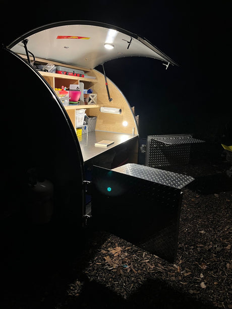 Inside a lit teardrop trailer galley at night, showing kitchen essentials and storage. Perfect for your next RV rental trip.