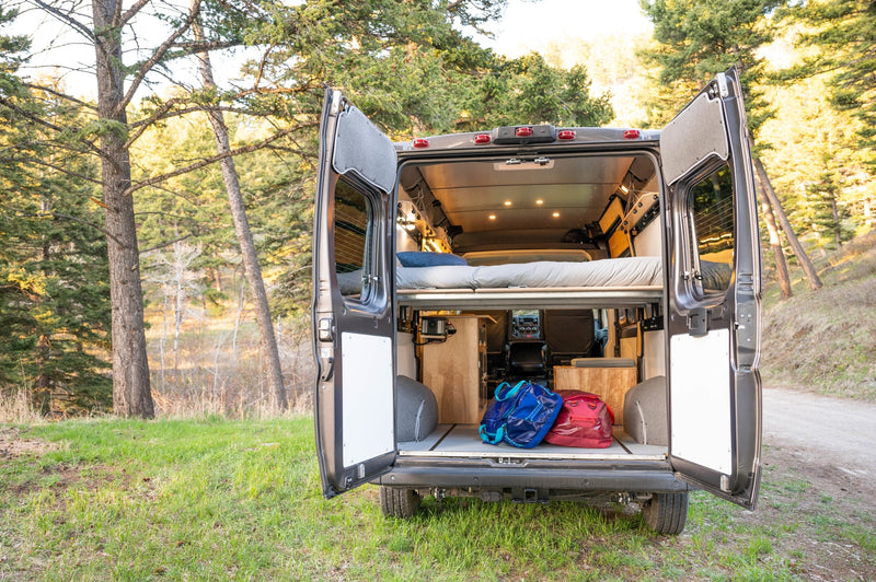 Open rv rental camper van rear view. Shows cozy bed, storage with bags, and illuminated interior, parked in a forest. — photo 3