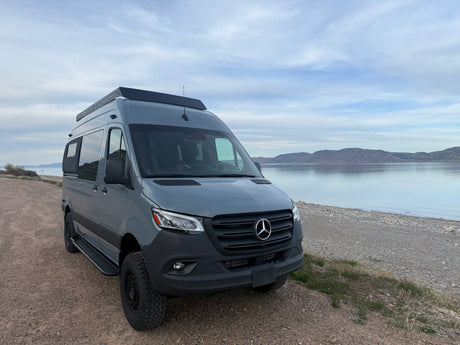 Experience freedom! Grey Mercedes-Benz Sprinter camper van, perfect RV rental, parked by a scenic lake with distant mountains.