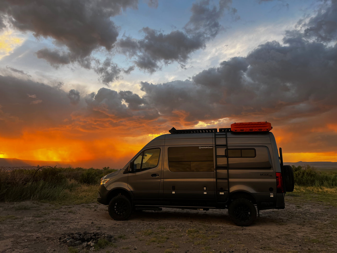 A grey camper van, ideal for an RV rental, is parked in a field under a dramatic sunset with vibrant orange light and dark clouds.
