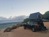 RV rental style: SUV with rooftop tent for beach camping. Chairs by the ocean, plane overhead.