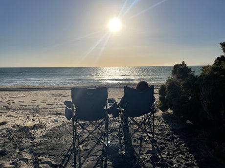 Unwind by the ocean with an RV rental adventure! Two chairs face the bright sun setting over the calm sea on a sandy beach.