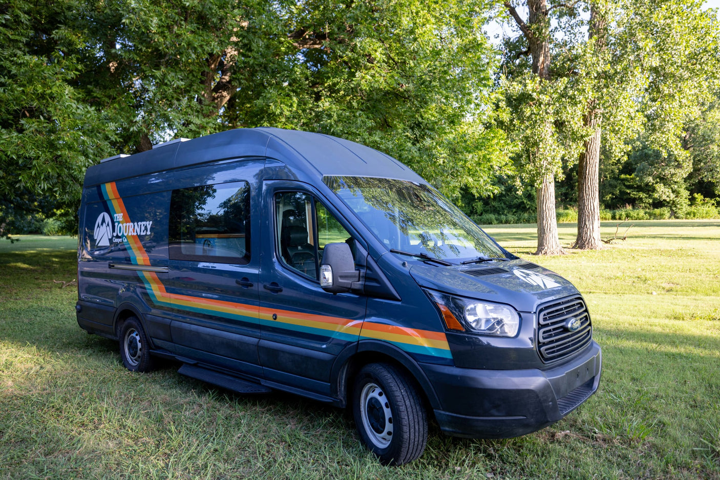  parked outdoors in a grassy area.|Explore with our "The Journey" camper van RV rental. This blue van with colorful stripes is parked on grass