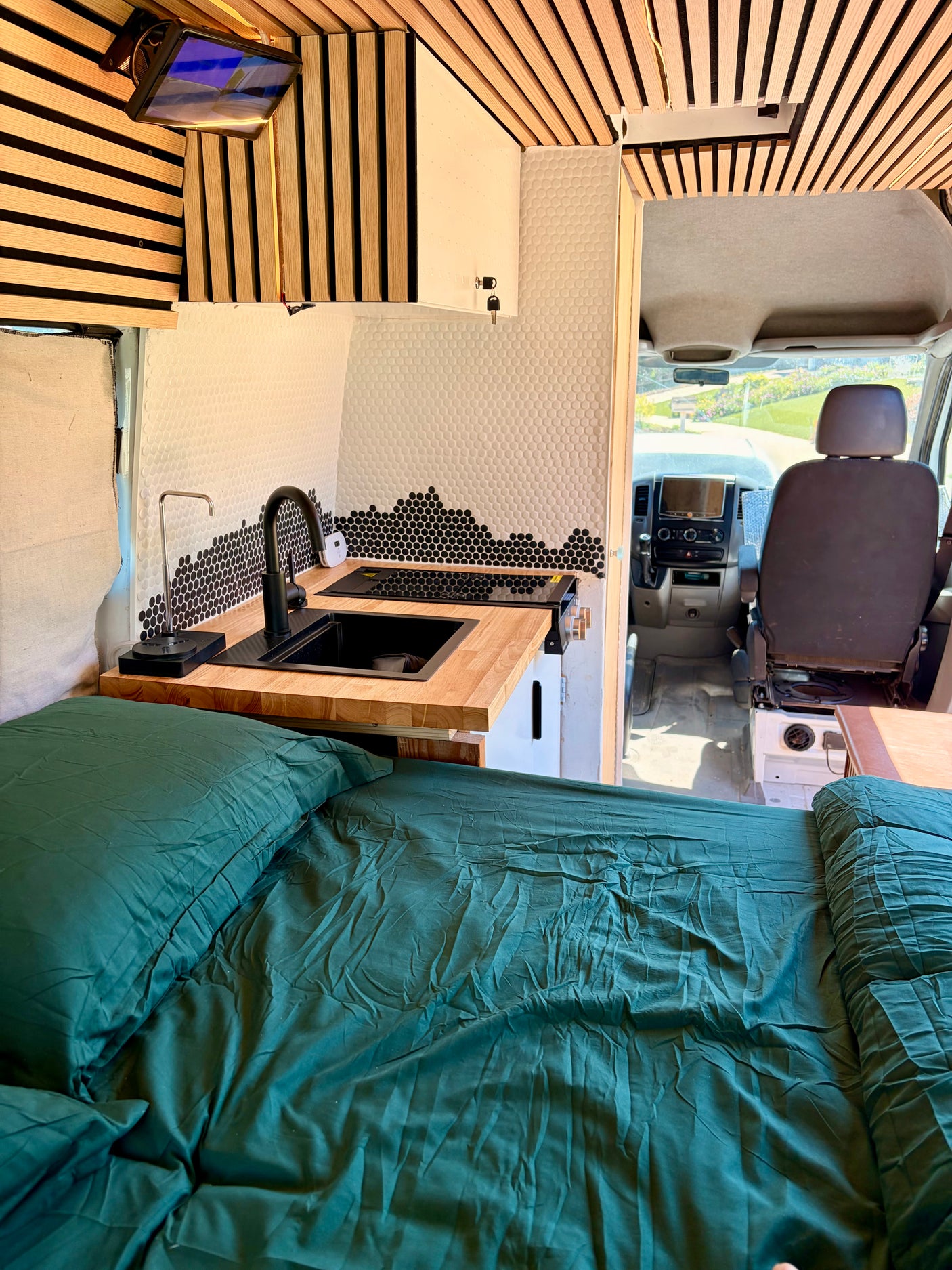Cozy dark green bed inside a modern camper van rv rental with a kitchen, wooden accents, and driver's area visible.