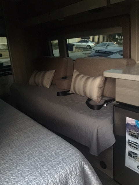 An RV rental's interior: a comfortable bed and seating area with a window view of parked vehicles outside.