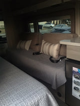 An RV rental's interior: a comfortable bed and seating area with a window view of parked vehicles outside.
