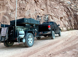 This RV rental setup is adventure-ready! A truck pulls a camping trailer with bikes on a dirt road by a towering canyon wall.