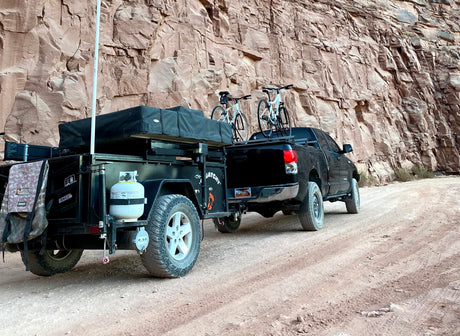 This RV rental setup is adventure-ready! A truck pulls a camping trailer with bikes on a dirt road by a towering canyon wall.