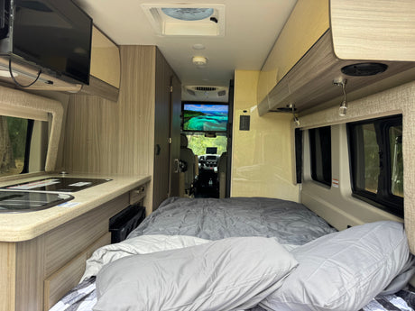 Inside a modern rv rental: comfortable bed, kitchen with sink & stovetop, TV showing a beautiful ocean scene, ready for adventure.