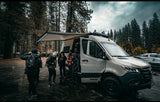 Group enjoys an RV rental van adventure in a damp forest. A light-colored Winnebago with an awning out, surrounded by tall trees.
