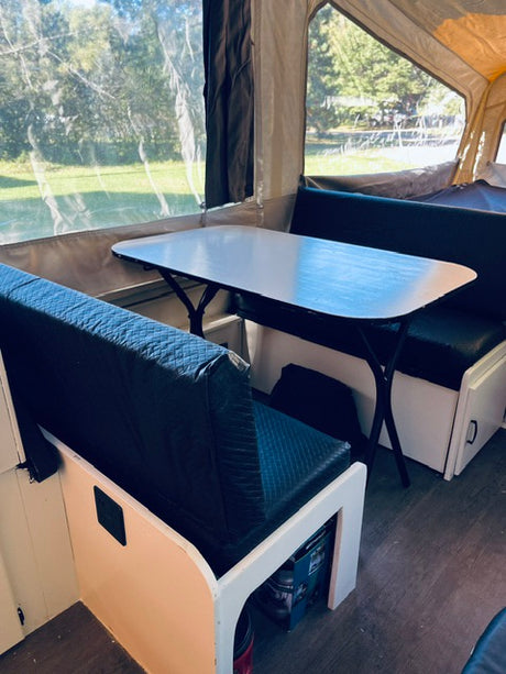 Dinette inside an rv rental pop-up camper with dark cushions, a white table, and clear window views of nature.
