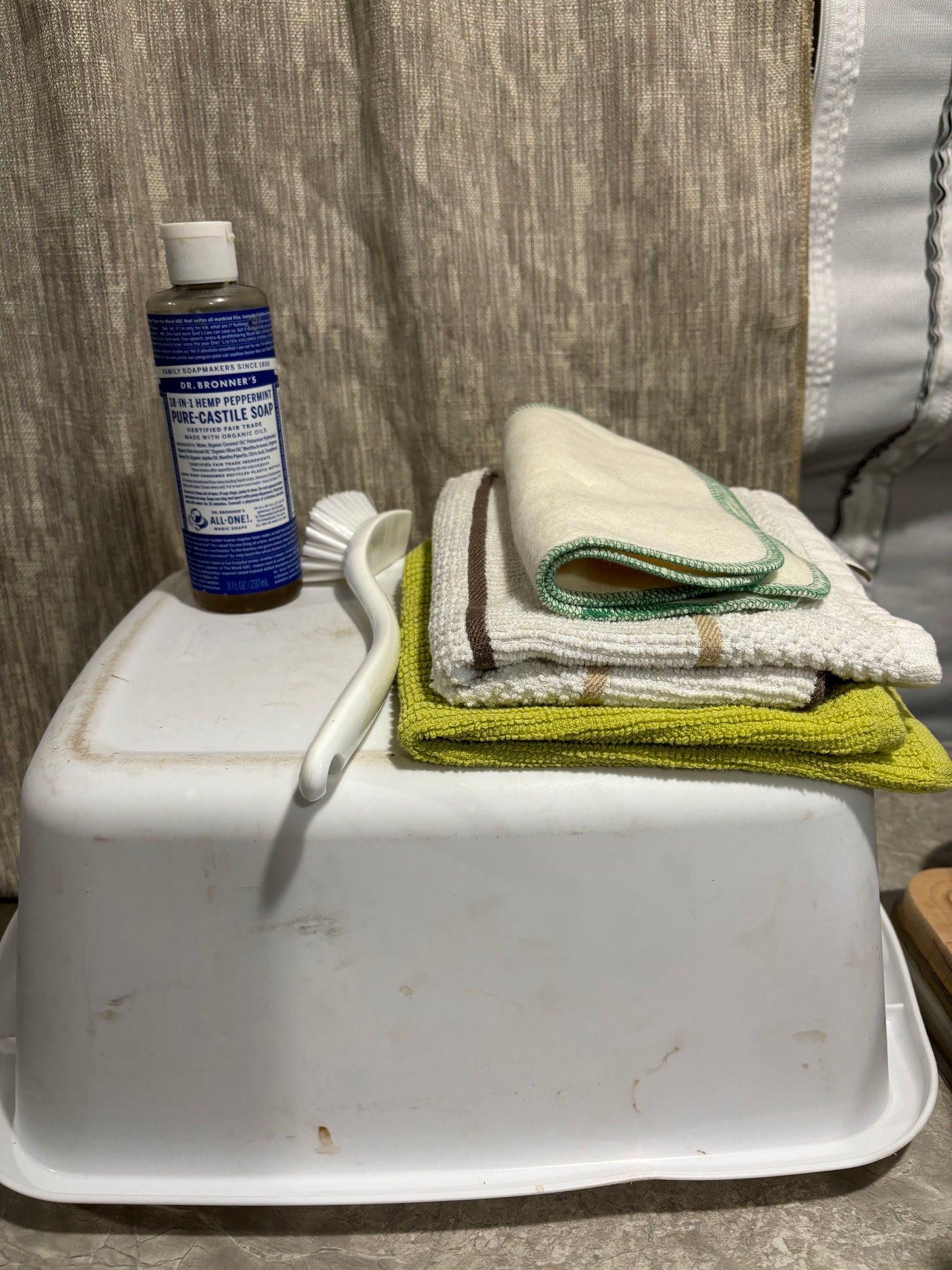 RV rental cleaning essentials: Dr. Bronner's soap, dish brush, & varied cloths on a white basin for compact living.