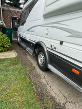 A Vista Cruiser RV rental, a white camper van, is parked on a paved driveway, showcasing its side features and utility access points.