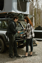 A couple cooks a meal using an RV rental's pull-out camp kitchen, attached to a white vehicle with a rooftop tent.
