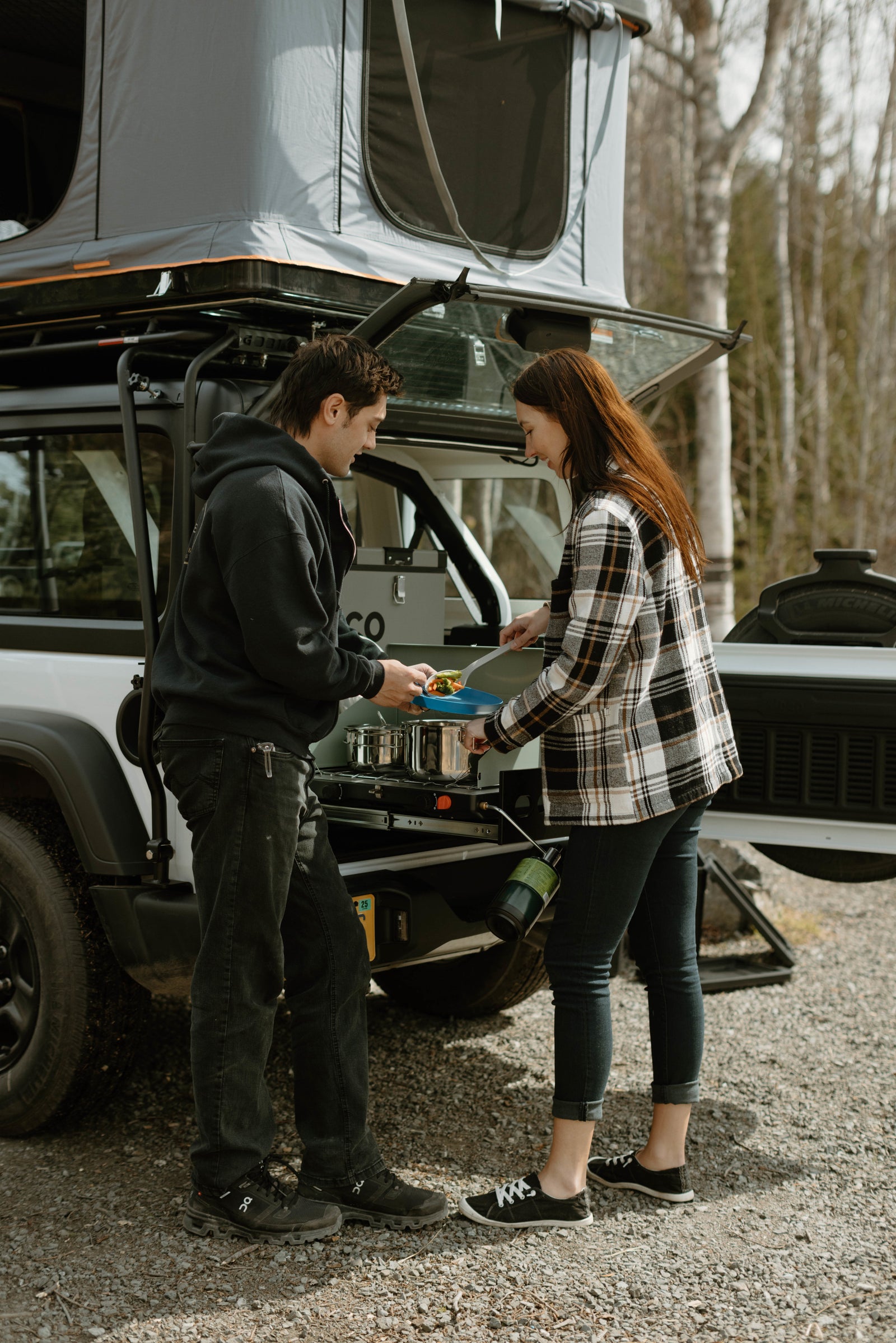 A couple cooks a meal using an RV rental's pull-out camp kitchen, attached to a white vehicle with a rooftop tent.