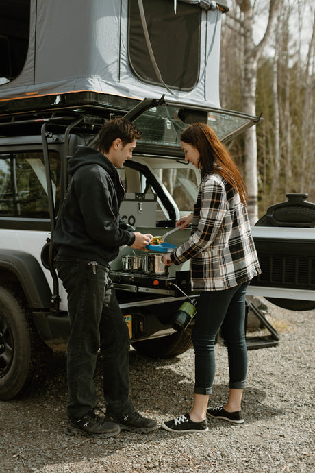 A couple cooks a meal using an RV rental's pull-out camp kitchen, attached to a white vehicle with a rooftop tent.