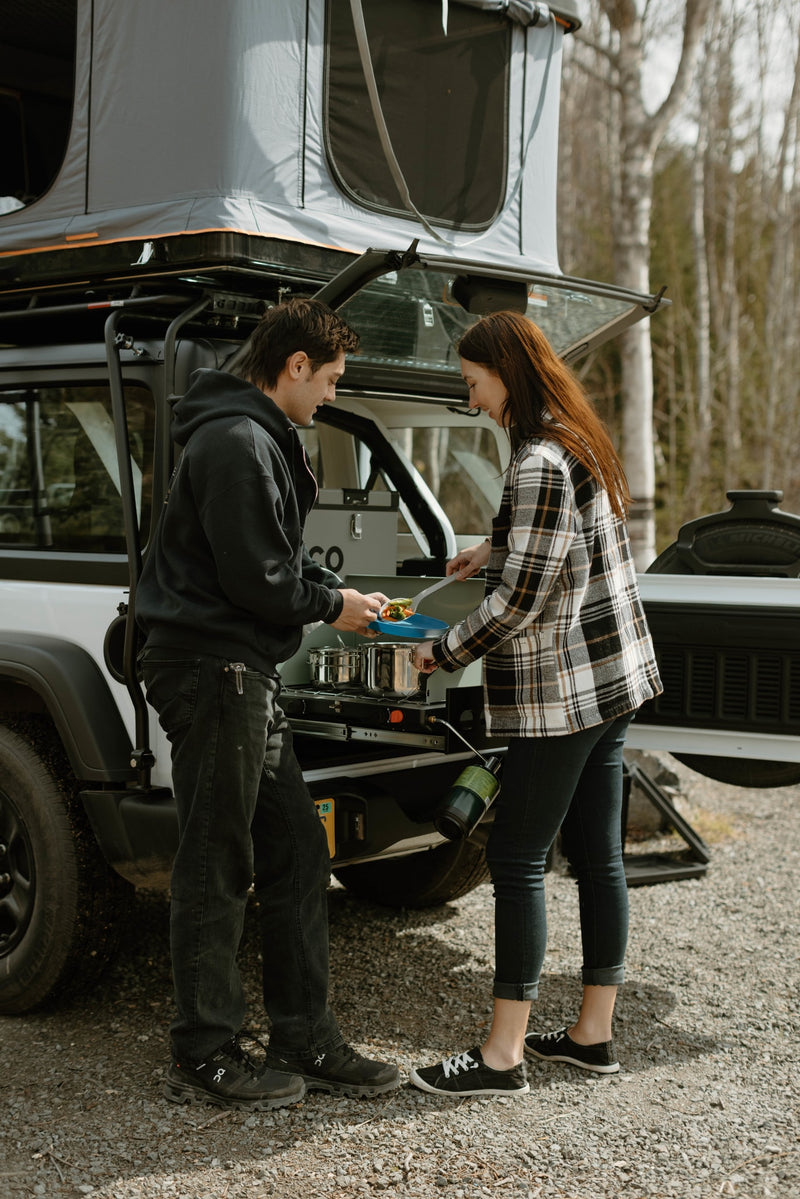 A couple cooks a meal using an RV rental's pull-out camp kitchen, attached to a white vehicle with a rooftop tent. — photo 12