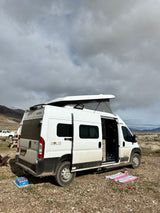 A white Solis camper van, likely an rv rental, is parked with its pop-top up, showing a camping setup in a vast outdoor landscape.