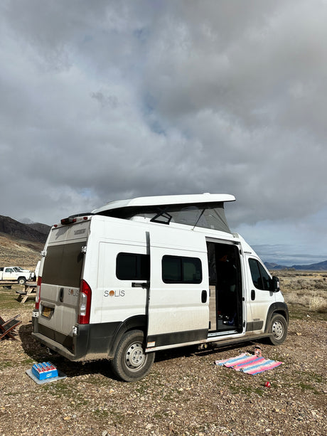 A white Solis camper van, likely an rv rental, is parked with its pop-top up, showing a camping setup in a vast outdoor landscape.