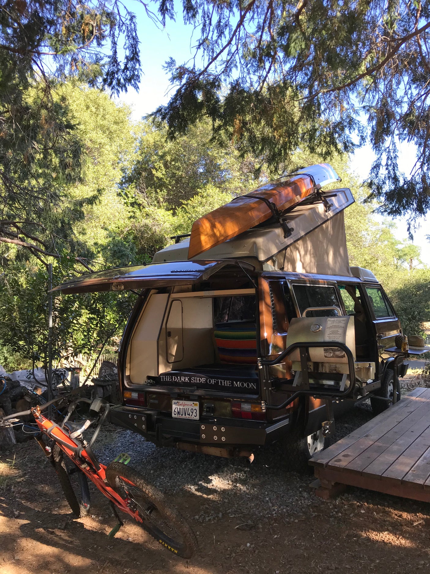 Brown VW camper van RV rental, equipped with a kayak, grill, and mountain bike, parked at a sunny outdoor campsite.