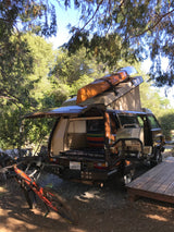 Brown VW camper van RV rental, equipped with a kayak, grill, and mountain bike, parked at a sunny outdoor campsite.
