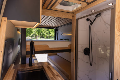 Inside an RV rental: a modern kitchen with wood counters, elevated bed, and tiled shower with black fixtures.