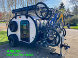 A white teardrop RV rental equipped with a 4-bike rack carrying multiple bicycles and a roof rack system for gear.