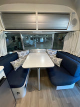 Inside an RV rental: a modern dining area with blue benches, a white table, and overhead storage. Other RVs reflect in the windows.