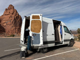 Adventure-ready RV rental van with climbing rope & backpack by towering red rock formations in a desert park.