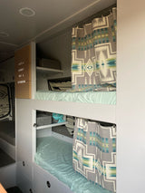 Inside a modern RV rental: light blue bunk beds with geometric patterned curtains, storage, and "Life's a Journey" sign.