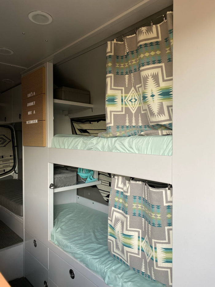 Inside a modern RV rental: light blue bunk beds with geometric patterned curtains, storage, and "Life's a Journey" sign.