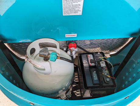 Inside an RV rental utility compartment, showing a white LP tank, a black battery, and a red battery disconnect switch.