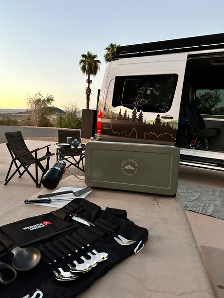 RV rental setup at sunset: A camper van with outdoor kitchen gear, chairs, & cutlery for desert adventures.
