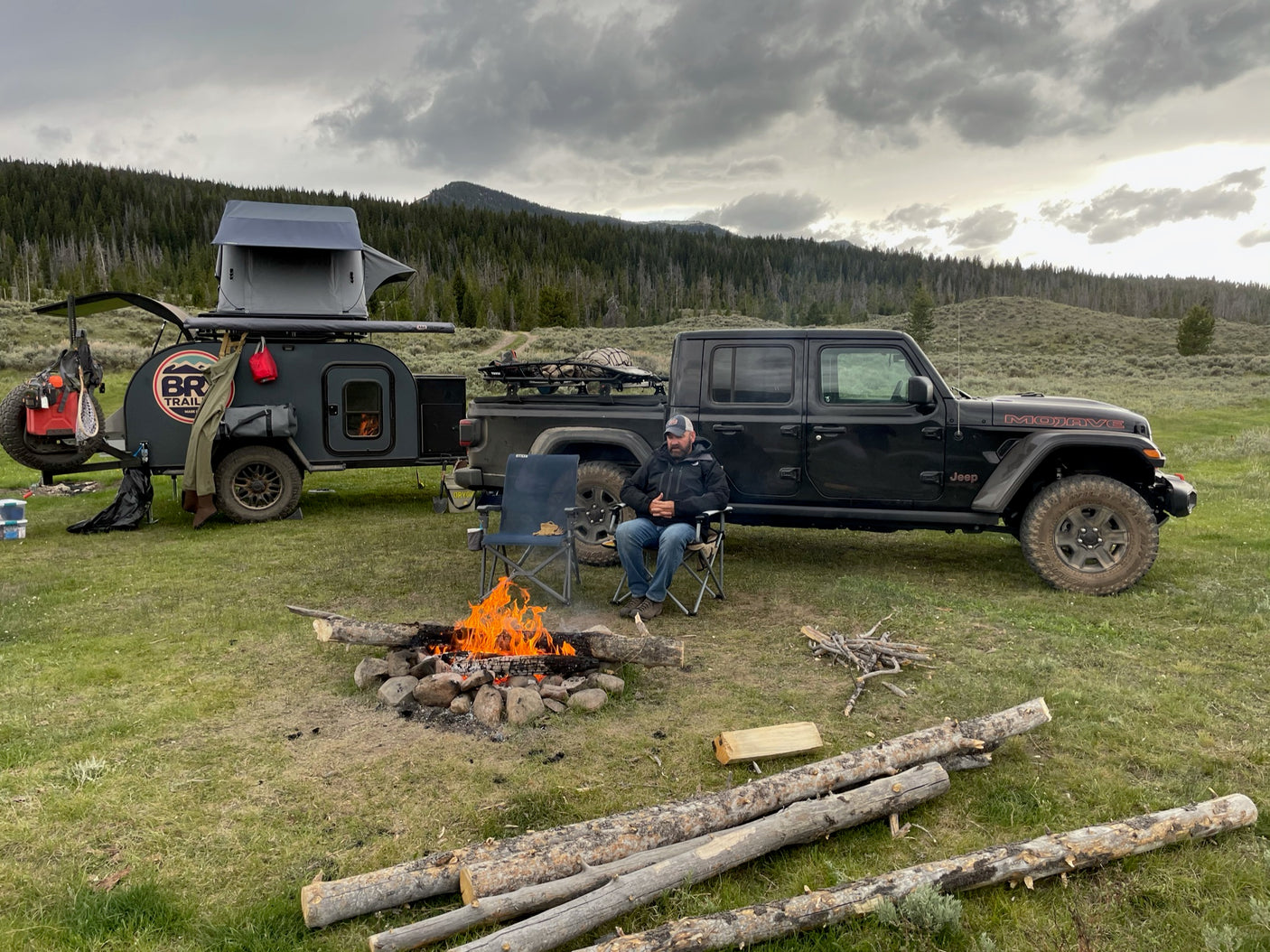 Off-road adventure: Man by campfire with a Jeep & teardrop camper. Explore with an RV rental like this!