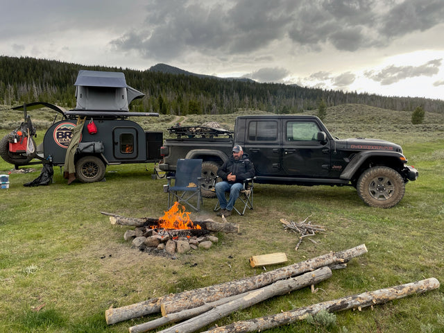 Off-road adventure: Man by campfire with a Jeep & teardrop camper. Explore with an RV rental like this!